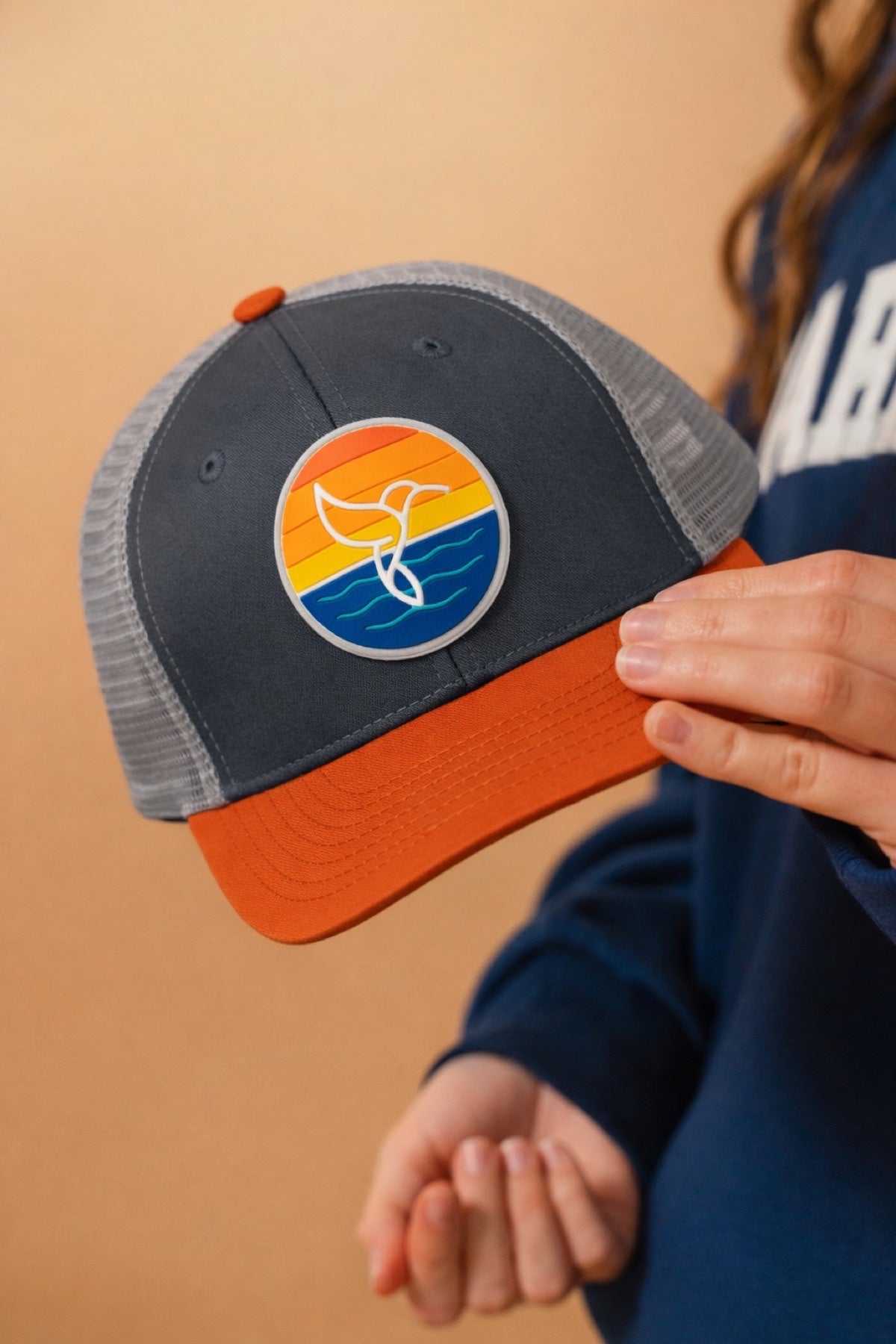 Hummingbird Sunrise Hat in Autumn Charcoal — model holding charcoal gray hat with orange brim and hummingbird sunrise patch | Warrior Sol cancer support accessories
