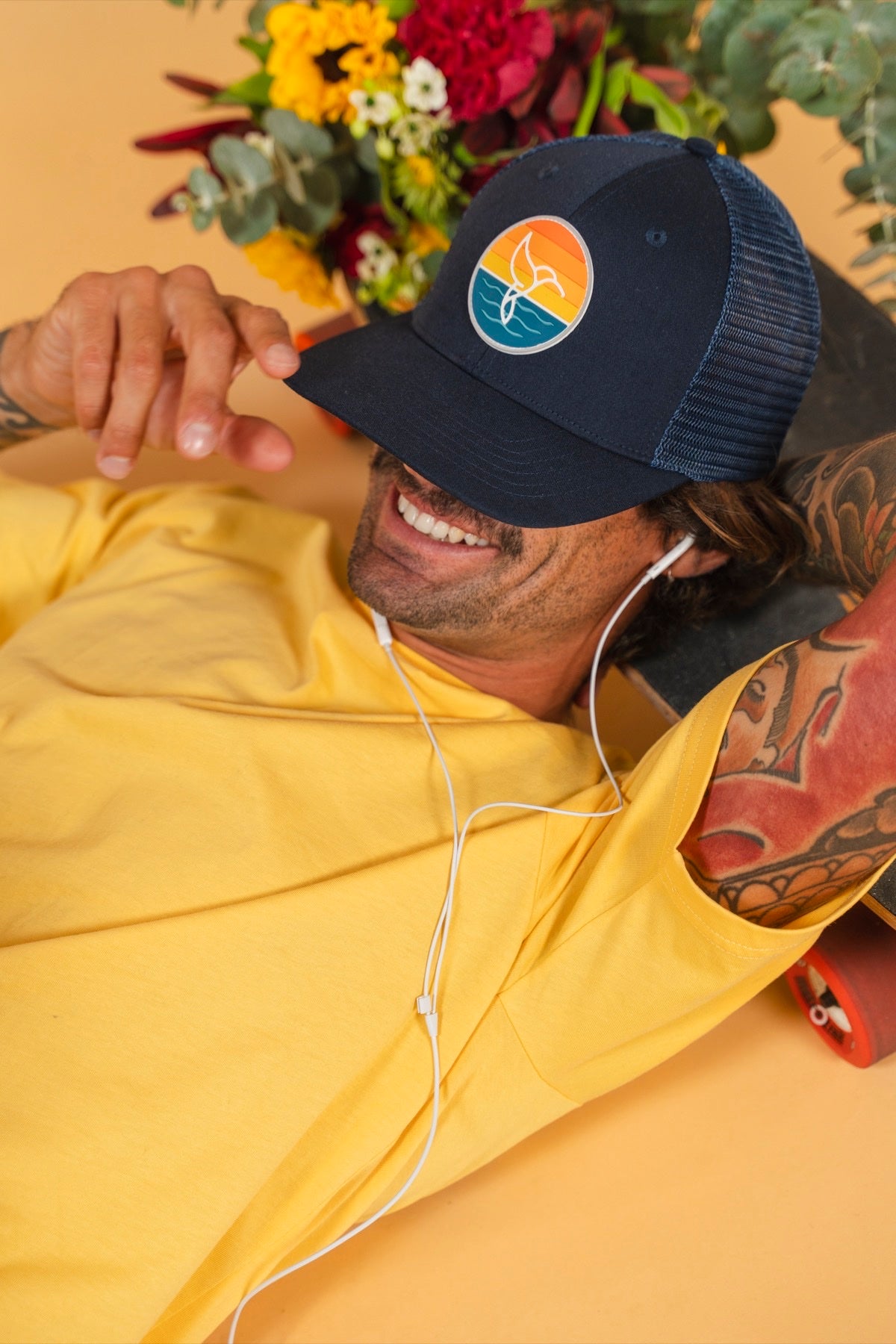 Hummingbird Sunrise Hat in Midnight — model wearing navy hat with hummingbird sunrise logo, yellow hoodie | Warrior Sol cancer support accessories