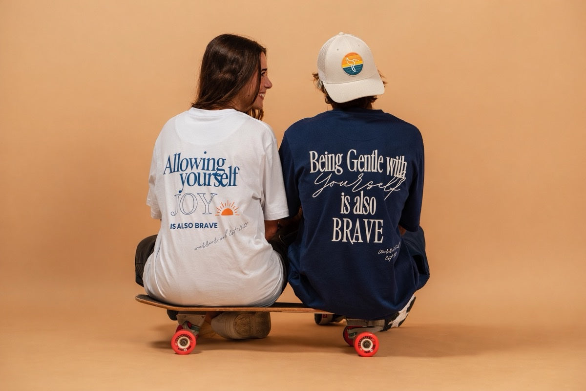 Hummingbird Sunrise Hat in Sand+Shell — two models on skateboard, cream hat with hummingbird sunrise logo visible | Warrior Sol cancer support accessories