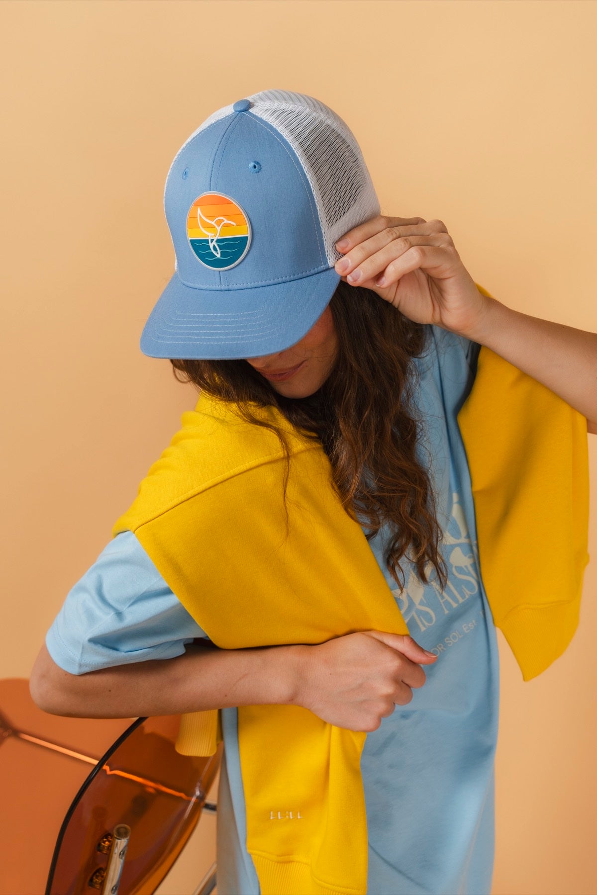 Hummingbird Sunrise Hat in Sky Blue — model holding sky blue hat with hummingbird sunrise logo, yellow hoodie | Warrior Sol cancer support accessories