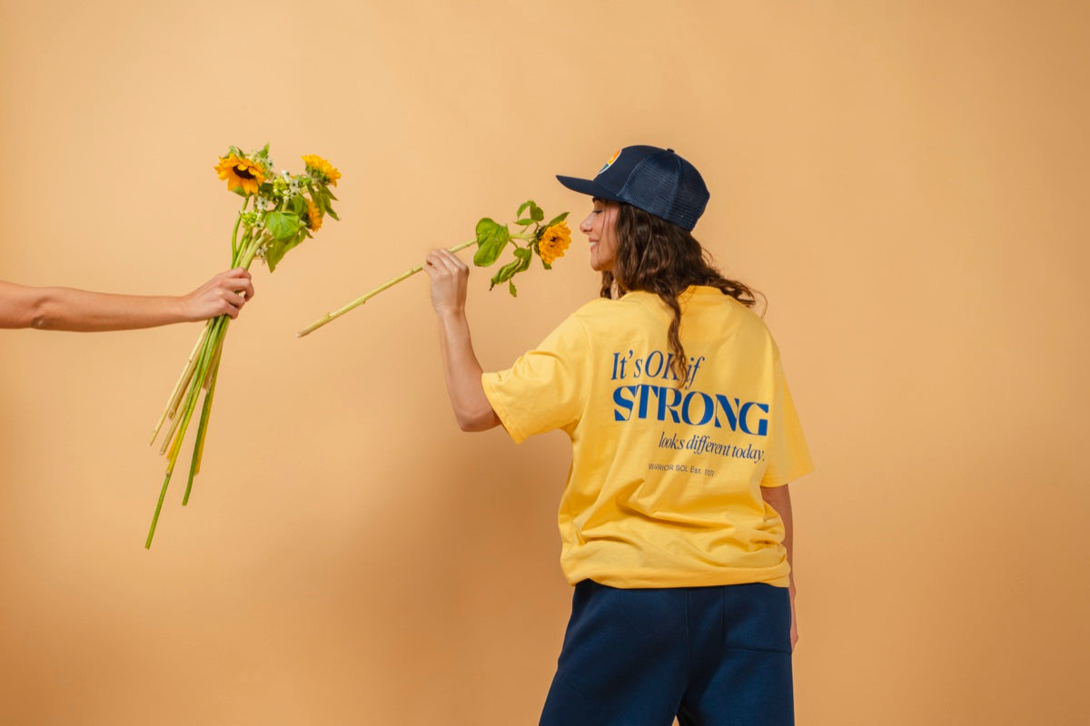 Warrior Sol Brave Series - Its OK if strong looks different today - female model with sunflowers wearing Sunshine Yellow t-shirt, warm gold campaign background