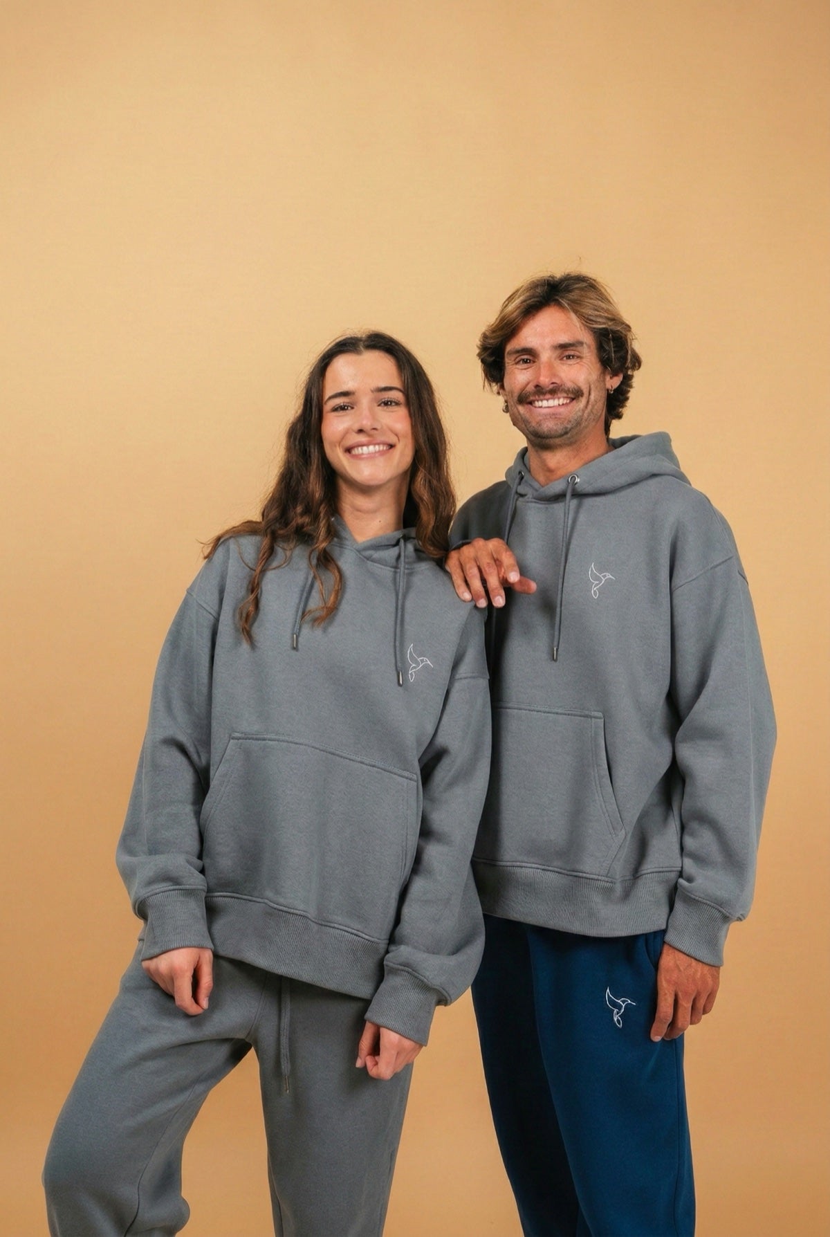 Warrior Sol Comfy Pullover Hoodie in Cloud Gray — campaign photo on gold background, two models wearing matching cloud gray hoodies with hummingbird logo